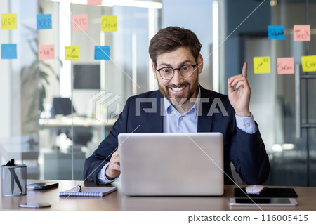 Businessman wearing suit working on laptop in modern office, smiling and raising finger as if having an idea. Sticky notes with various text on glass wall in background. Businessman wearing suit working on laptop in modern office, smiling and raising finger as if having an idea. Sticky notes with various text on glass wall in background. 116005415