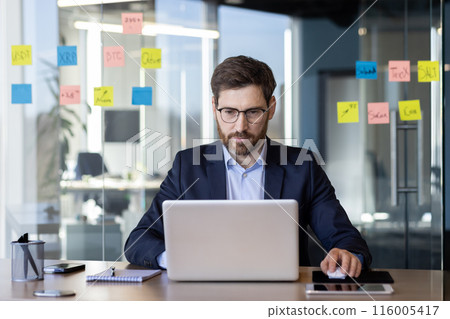 Businessman in formal suit focusing on laptop in contemporary office. Sticky notes with various information on glass wall. Corporate environment, productive work, technology usage, and business Businessman in formal suit focusing on laptop in contemporary office. Sticky notes with various information on glass wall. Corporate environment, productive work, technology usage, and business 116005417