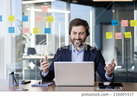 Smiling businessman using laptop and headset for virtual meeting in modern office setting. Professional office environment with sticky notes on glass wall in background Smiling businessman using laptop and headset for virtual meeting in modern office setting. Professional office environment with sticky notes on glass wall in background 116005437