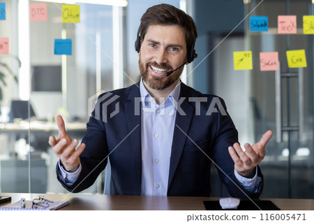 Smiling businessman wearing suit and headset engaged in virtual meeting in modern office. Confident professional communicating remotely using video conferencing technology, interactive work 116005471