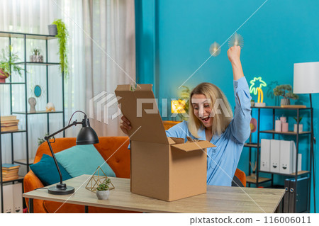 Happy blond businesswoman unpacking opening delivery parcel box purchase sits at desk in apartment 116006011