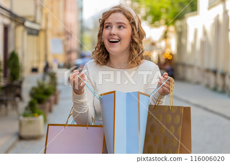 Happy young woman shopaholic consumer after shopping sale with full bags walking in city town street 116006020