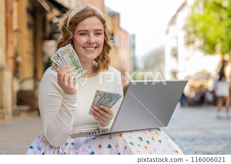 Happy rich winner girl using laptop victory counting cash money dollar banknotes sitting on street Happy rich winner girl using laptop victory counting cash money dollar banknotes sitting on street 116006021