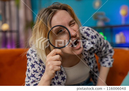 Woman holding magnifying glass near face looking into camera with big zoomed funny eye analyzing 116006048