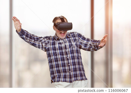 Portrait of excited old man in headset with his arms spreaded. Blurred windows background. 116006067