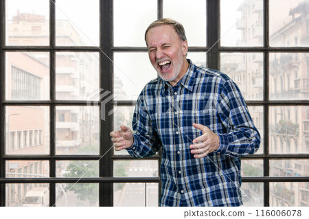 Handsome aged senior man smiling and laughing hard out loud because of funny joke. Standing indoors in office. Handsome aged senior man smiling and laughing hard out loud because of funny joke. Standing indoors in office. 116006078