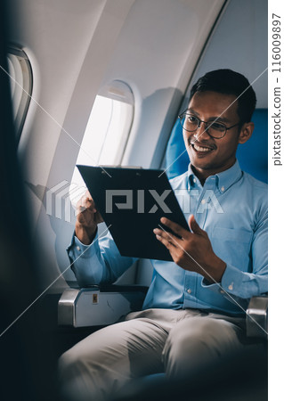Attractive Asian male passenger of airplane sitting in comfortable seat while working laptop and tablet with mock up area using wireless connection. Travel in style, work with grace. 116009897