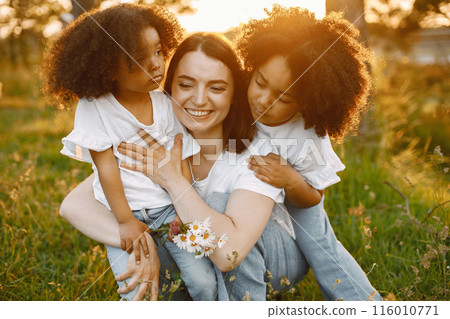 Photo of caucasian mother and two her african american daughters embracing together outdoors. Girls has black curly hair. Mother and daughers wearing white t-shirts. Photo of caucasian mother and two her african american daughters embracing together outdoors. Girls has black curly hair. Mother and daughers wearing white t-shirts. 116010771