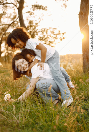 Photo of caucasian mother and two her african american daughters embracing together outdoors. Girls has black curly hair. Mother and daughers wearing white t-shirts. Photo of caucasian mother and two her african american daughters embracing together outdoors. Girls has black curly hair. Mother and daughers wearing white t-shirts. 116010775