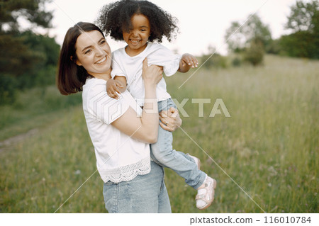 Photo of caucasian mother and her african american daughter embracing together outdoors. Girl has black curly hair. Mother and daugher wearing white t-shirts. 116010784