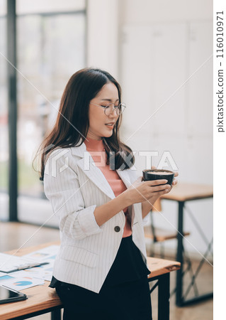 Beautiful young smiling Asian businesswoman working on laptop and drinking coffee, Asia businesswoman working document finance and calculator in her office. 116010971