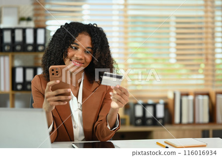Smiling African American female worker holding mobile phone and credit card for financial transaction 116016934