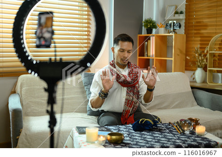 Fortune teller casting a spell near burning candles while forecasting future online with smartphone in foreground Fortune teller casting a spell near burning candles while forecasting future online with smartphone in foreground 116016967