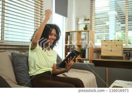 Cheerful young woman looking at digital tablet computer celebrating victory on couch, getting great good news 116016971