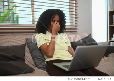 African American woman sitting on sofa massaging nose bridge, feeling eye strain during working at home 116016973