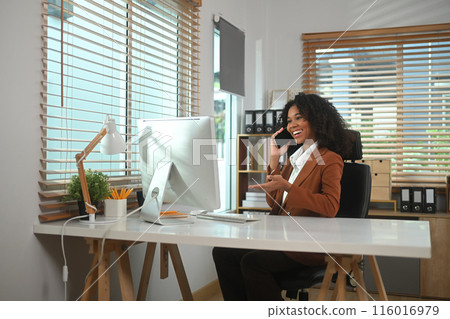 Cheerful African American businesswoman talking on phone and smiling while sitting at her office Cheerful African American businesswoman talking on phone and smiling while sitting at her office 116016979