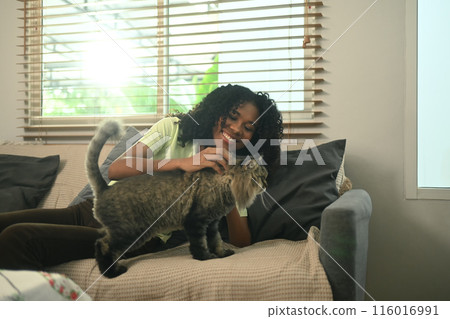Happy young woman with curly hair relaxing on a cozy sofa while lovingly petting her fluffy cat Happy young woman with curly hair relaxing on a cozy sofa while lovingly petting her fluffy cat 116016991