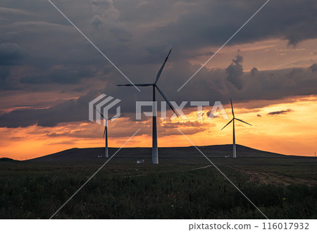 Multiple wind turbines silhouetted against a vibrant sunset sky. The turbines stand tall against the backdrop of orange and red clouds. 116017932