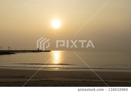 Orange hues paint the sky as the sun dips below the horizon, casting a warm glow on the ocean waves lapping at the sandy shore 116018960