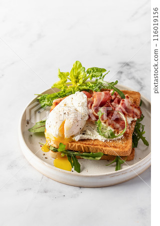 Toasts sandwich with poached egg, arugula, bacon and cream cheese on white plate marble background. Isolated, text space Toasts sandwich with poached egg, arugula, bacon and cream cheese on white plate marble background. Isolated, text space 116019156