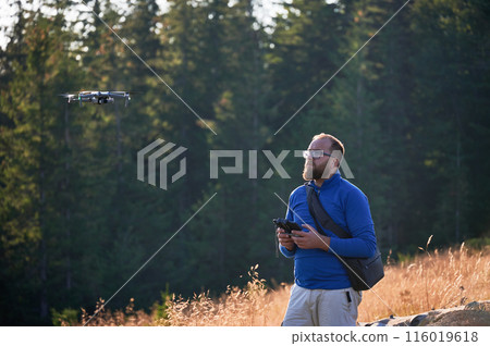 Professional photographer taking photos from drone. Young man taking aerial photos in mountains. Man controlling his drone on background of mountain forest. Professional photographer taking photos from drone. Young man taking aerial photos in mountains. Man controlling his drone on background of mountain forest. 116019618