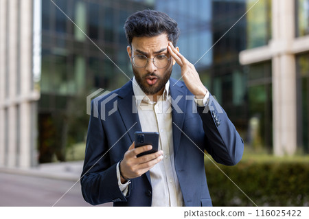Shocked businessman in blue suit looking at smartphone with surprised expression outside modern office building, feeling shocked, stressed, and concerned while reading unexpected news or error. 116025422