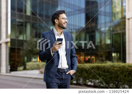 Confident young businessman in suit holding smartphone and smiling while standing outdoors near modern office building. Professional attitude, business success, and positive outlook convey success Confident young businessman in suit holding smartphone and smiling while standing outdoors near modern office building. Professional attitude, business success, and positive outlook convey success 116025424