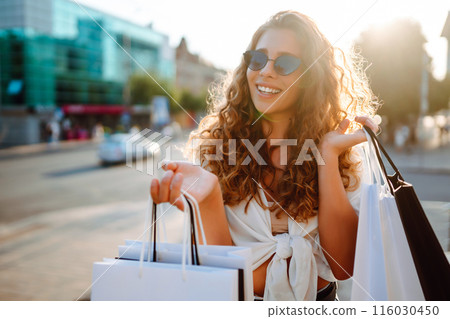 Stylish woman with shopping bags walks through city street. Consumerism, shopping, lifestyle concept Stylish woman with shopping bags walks through city street. Consumerism, shopping, lifestyle concept 116030450