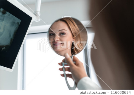 In the dental chair, the patient is holding a mirror, happy and grateful for the high-quality work of the dentist who made her smile perfect. A patient in a chair, holding a mirror, enjoys every 116030654