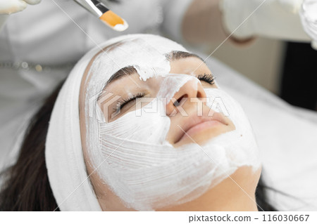 A woman is receiving a professional facial treatment at a spa, applying a white cream mask to rejuvenate her skin and achieve a radiant, youthful look. The atmosphere is tranquil and relaxing 116030667