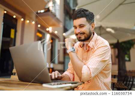 Smiling young man working in cafe on street with laptop and phone. Freelance business concept. Smiling young man working in cafe on street with laptop and phone. Freelance business concept. 116030734