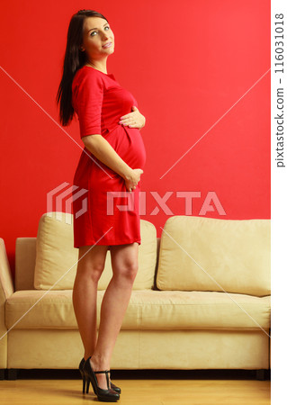 pregnant woman in red dress touching her belly at home pregnant woman in red dress touching her belly at home 116031018