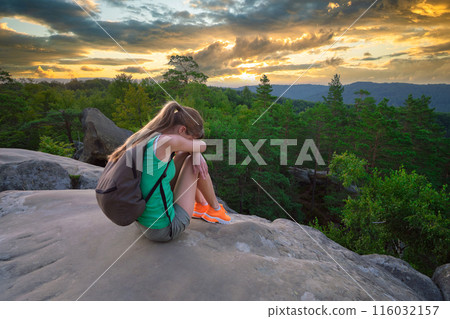 Lonely young woman sitting depressed on hillside trail on warm summer evening. Lost female hiker crying on rocky cliff on wilderness path. Depression and loneliness concept 116032157