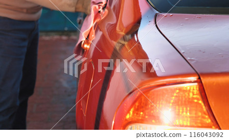 A man thoroughly washes an orange car with a pink rag. The shiny surface of the car and the water droplets on it indicate a recent wash, which shows the care and attention given to the car. 116043092
