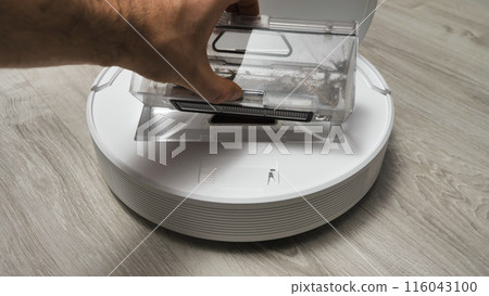 A man's hand removes a container of debris from the robot vacuum cleaner. A robot vacuum cleaner on a light-colored floor designed for automatic cleaning. 116043100