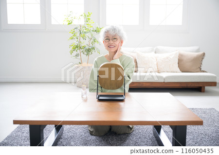 Front view of a senior (elderly) Japanese woman taking care of her skin (skin care) looking in the mirror in a bright living room Camera Front view of a senior (elderly) Japanese woman taking care of her skin (skin care) looking in the mirror in a bright living room Camera 116050435