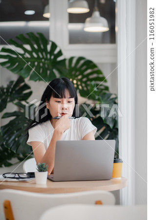 Young woman working on a laptop in a modern office with green plants in the background, contemplating ideas, and holding a pen. Young woman working on a laptop in a modern office with green plants in the background, contemplating ideas, and holding a pen. 116051982
