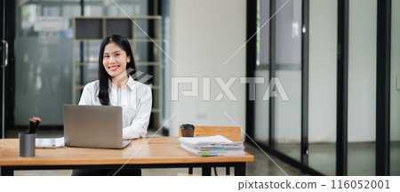 Young professional woman working at a modern office desk with a laptop and documents, smiling confidently. Young professional woman working at a modern office desk with a laptop and documents, smiling confidently. 116052001