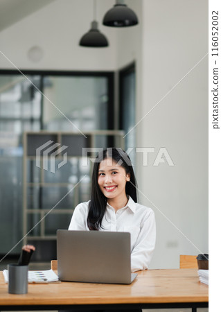 Confident young woman smiling while working on a laptop in a modern office setting with minimalist design and natural lighting. 116052002