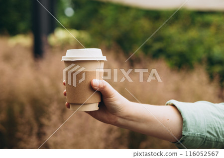 Coffee to go at autumn park. Food, rest, Take away concept. Place logo on mug, mockup. Woman hand holding brown paper cup on park background 116052567