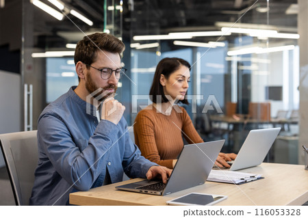 Two focused professionals working together at a desk with laptops in a contemporary office setting, conveying collaboration and teamwork. Two focused professionals working together at a desk with laptops in a contemporary office setting, conveying collaboration and teamwork. 116053328