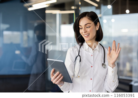 Young female doctor using tablet for video call in modern office. Professional in white coat with stethoscope having virtual consultation, connecting with patients online, waving at screen 116053391