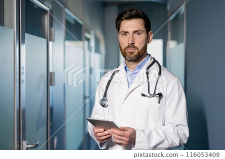 Confident doctor wearing white coat with stethoscope standing in clinic hallway holding tablet. Medical professional ready for patient consultation. Healthcare worker in modern medical facility 116053409