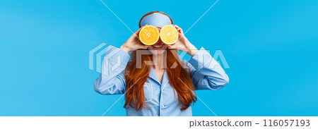Carefree pretty and cute redhead woman in nightwear, sleep mask, holding two slices of orange over eyes and smiling, fool around in morning, standing blue background joyful 116057193