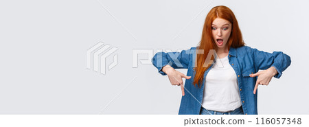 Waist-up portrait impressed, curious and astonished redhead cheerful girl in denim shirt, drop jaw, looking and pointing down starled speechless, standing white background amused 116057348