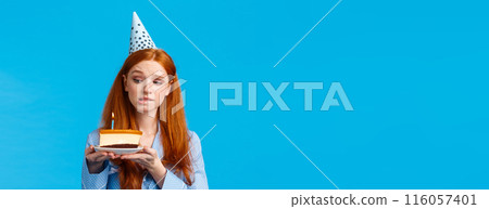 Attractive caucasian redhead woman in birthday cap, holding delicious, tasty b-day cake with lit candle, biting lip want eat dessert but worry about calories during diet, stand blue background Attractive caucasian redhead woman in birthday cap, holding delicious, tasty b-day cake with lit candle, biting lip want eat dessert but worry about calories during diet, stand blue background 116057401