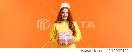Cute redhead european woman prepare gift, wrapped present and giving it on christmas holidays, wearing winter beanie, smiling joyfully as congratulating someone, orange background 116057503