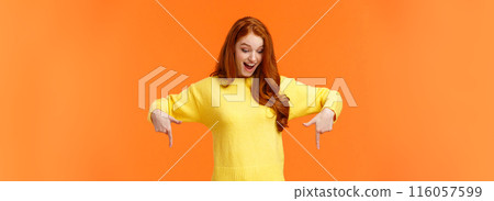 Wondered and excited, amused cute redhead woman in yellow sweater pointing fingers down, look bottom and smiling, check-out product promo, winter holiday event, orange background 116057599