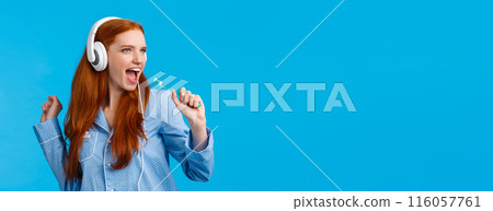 Sassy good-looking redhead female in pyjama having fun, listening music in big white headphones, singing into toothbrush as brushing teeth, getting ready go out, standing blue background Sassy good-looking redhead female in pyjama having fun, listening music in big white headphones, singing into toothbrush as brushing teeth, getting ready go out, standing blue background 116057761