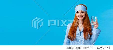 Waist-up shot glamour redhead woman taking care health, eating healthy, drinking water, wearing sleep mask and nightwear, holding glass and smiling energized, standing blue background 116057863